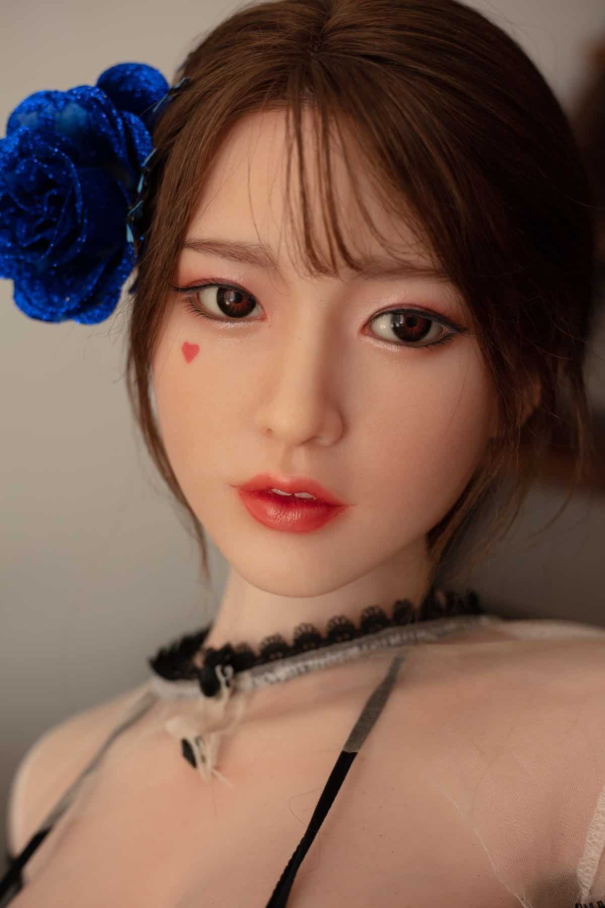 Living doll with realistic features and a blue floral hair accessory, wearing a sheer top with lace details.