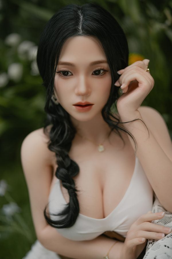Beautiful realistic female doll with black hair, wearing a white dress, posing outdoors among greenery.