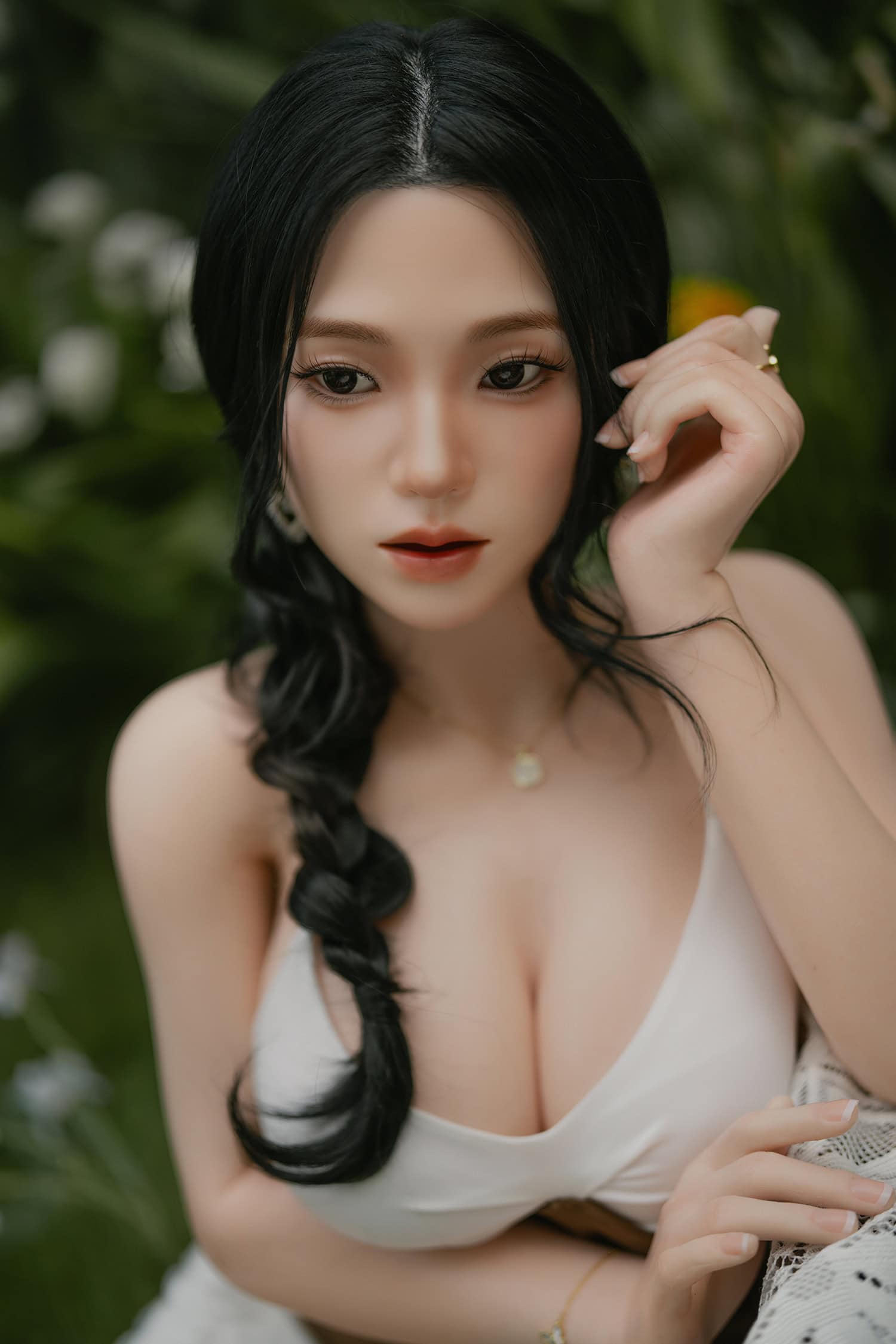 Beautiful realistic female doll with black hair, wearing a white dress, posing outdoors among greenery.