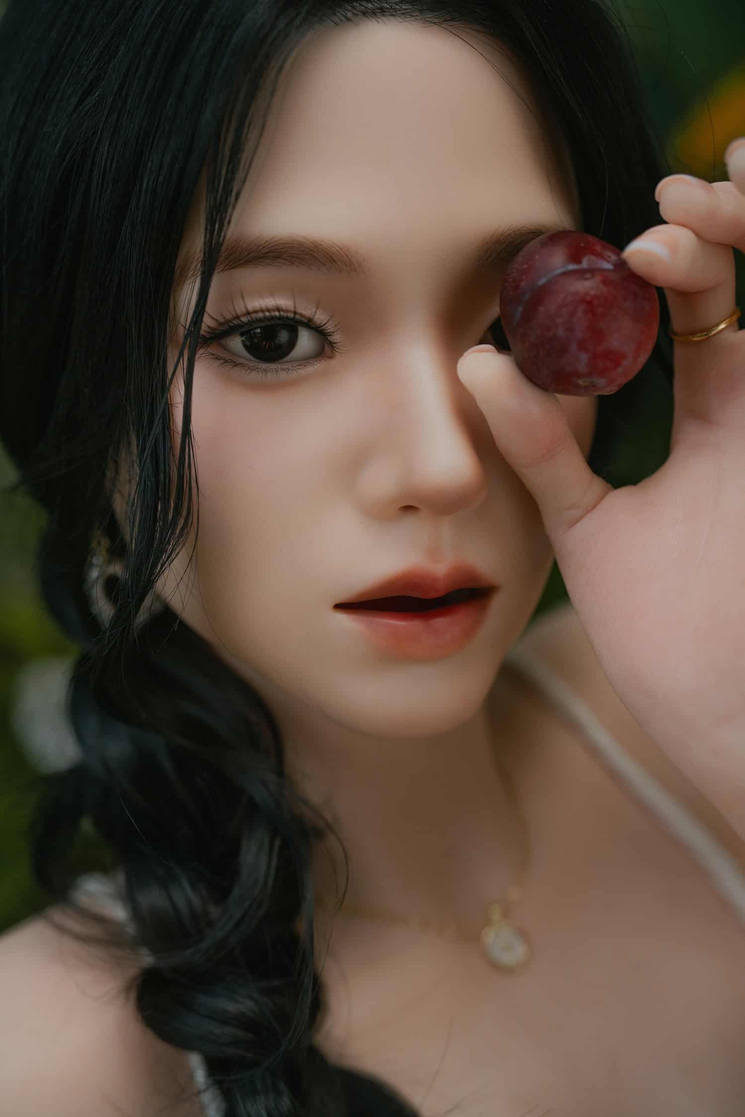Silicone doll with black hair holding a red apple near her eye, outdoor portrait.