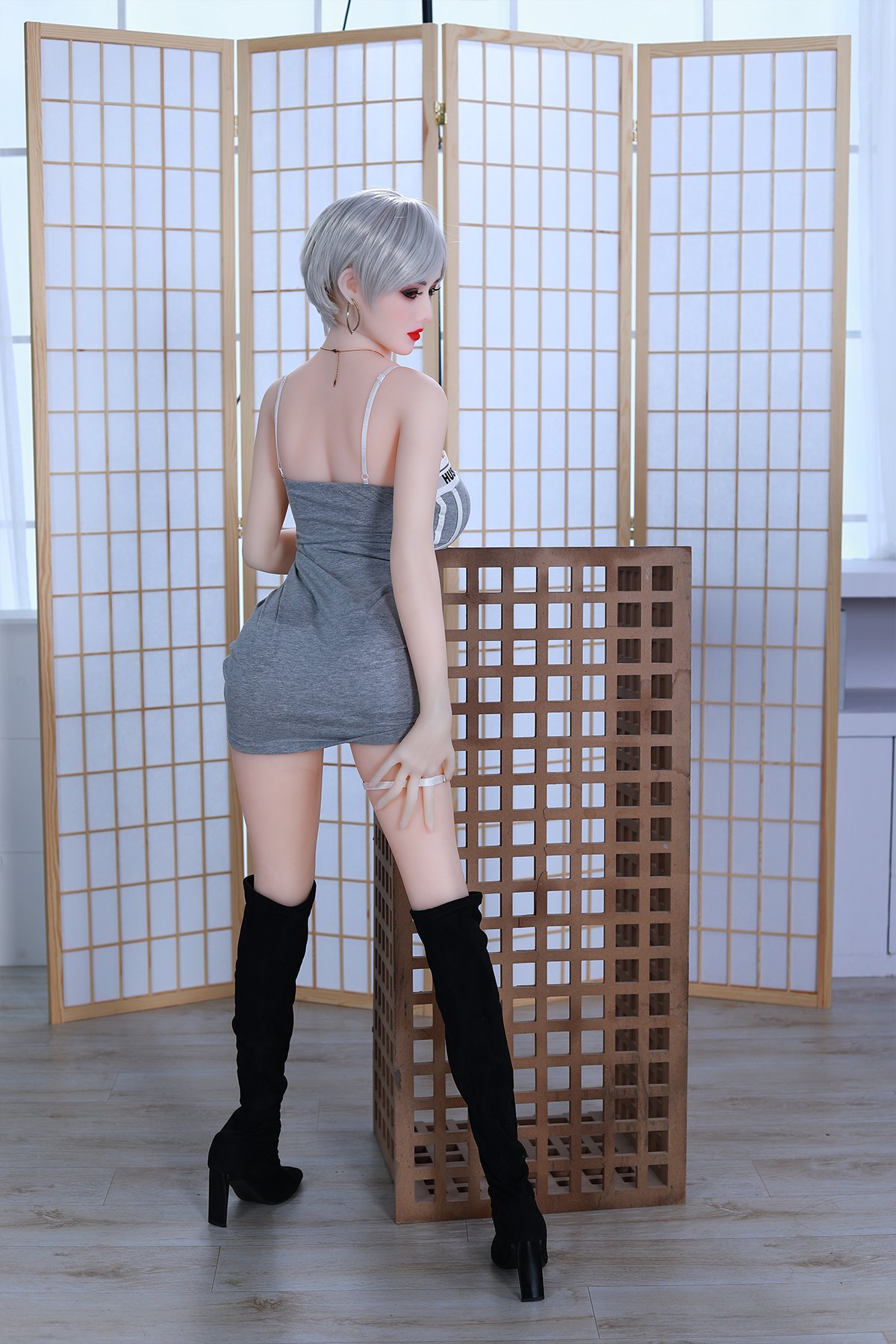 Silicone sex doll with short gray hair wearing a gray mini dress and black thigh-high boots in a room with wooden room dividers.