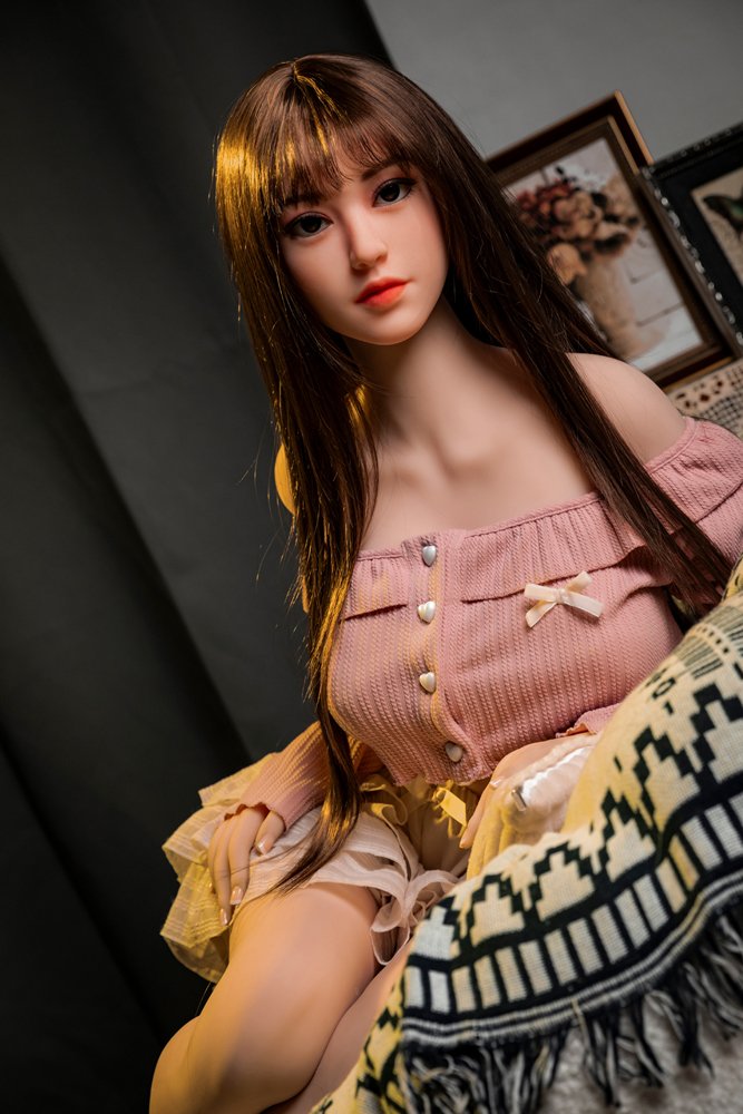 Realistic love doll sitting on bed with detailed features and lifelike appearance.