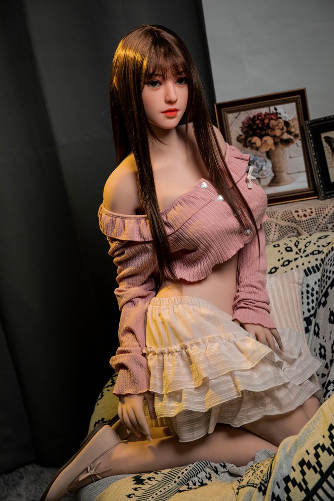 Realistic love doll with long brown hair and lifelike features, dressed in pink and beige, posed on cozy patterned sofa.