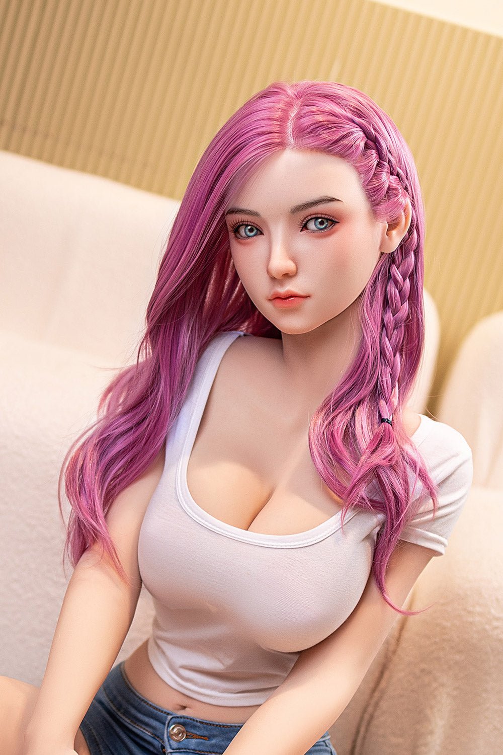 Silicone doll with pink braided hair, blue eyes, and white tank top, sitting on beige sofa for realistic intimacy.