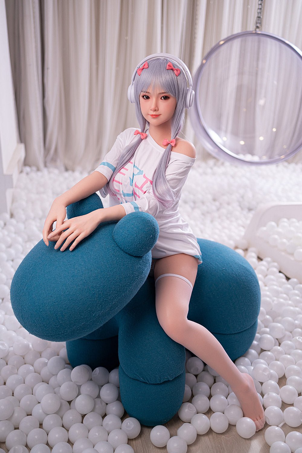 Cute anime-style sex doll in a playful pose with a bear-shaped chair, surrounded by white balls and soft lighting.