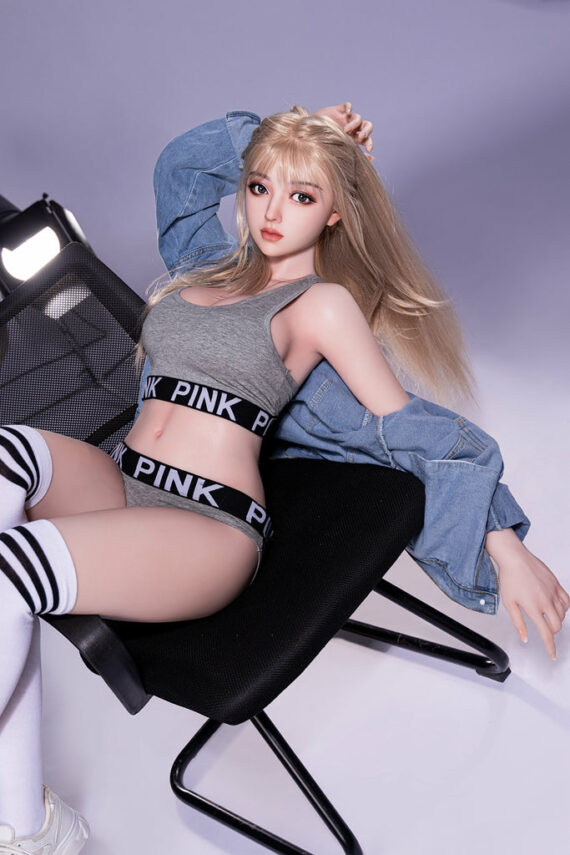 Realistic sex doll female with blonde hair in casual sportswear, posing on modern chair, sexy lifestyle model image.